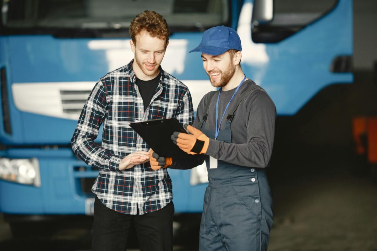 Two men discussing truck repairs in a garage: one in mechanic uniform, the other in a plaid shirt.